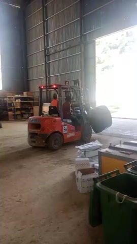 A forklift is being used inside a warehouse to move a large, dark, circular object. The forklift is orange and has a person operating it. The warehouse appears to have a metal corrugated exterior and a wide opening leading to the outside, where trees and bright daylight are visible. Boxes and other items are stacked on shelves and on the floor within the warehouse. Two green bins are also visible in the foreground.