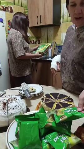 The video shows two women in a kitchen. The woman in the foreground is wearing a brown, textured shirt and has her right arm in a cast. She is looking down and appears to be placing items on a table. In front of her on the table are two cakes, one white and one with chocolate topping, and several green packages of what appear to be cookies or snacks. There is also a plate with a slice of cake and a serving utensil. Behind her, a woman with dark hair wearing a leopard-print top is standing by a counter and a sink, holding a green box. The kitchen has wooden cabinets and a green and yellow patterned backsplash. There is also a refrigerator with a calendar visible on the side. The lighting suggests it is daytime.