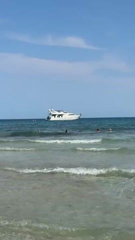 A white yacht floats on a calm blue ocean under a partly cloudy sky. The water near the shore has small, gentle waves breaking. Several people are visible in the water, appearing to be swimming or wading. The scene is bright and appears to be a sunny day at the beach.