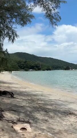 A static shot of a tropical beach scene. The foreground consists of sand and shadows from unseen trees. In the middle ground, a calm, turquoise ocean meets a sandy beach. The background features lush, green, tree-covered hills under a bright blue sky with scattered white clouds. A single, small, dark-colored rock formation is visible in the water on the right side of the frame. The overall impression is peaceful and serene.