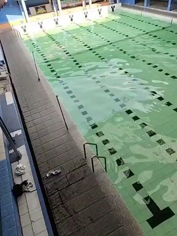 This is an aerial video of an empty swimming pool. The pool is rectangular and has lane markers on the bottom. The water is green and clear. There are blue and yellow banners hanging above the pool. There are also some chairs and tables visible. The pool appears to be indoors, as there is a roof above it. The video is shaky and the lighting is not very good. There are no people visible in the video.
