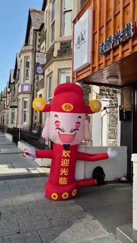 The video shows a large inflatable figure standing in front of a restaurant. The figure is red and white, with a smiling face and wearing a traditional Chinese hat. The restaurant behind it has a wooden exterior and a sign with Chinese characters. Pedestrians are walking on the sidewalk. One man stops to interact with the inflatable figure. The sky is clear and blue. The scene appears to be a sunny day on a city street.