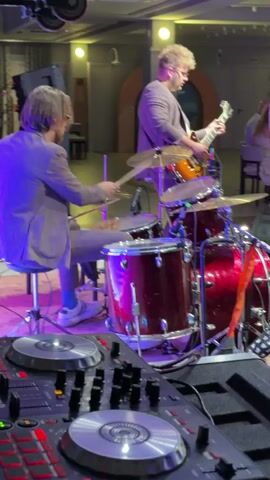 The video captures a band performing on a stage, bathed in purple and white lighting. In the foreground, a DJ setup is visible, with illuminated buttons and turntables. To the left, a drummer is seated, playing a red drum set, while a guitarist stands to the right, playing an electric guitar. Another person, likely a band member, is partially visible in the foreground, wearing a dark suit and facing away from the camera. In the background, audience members are seated at tables. The scene suggests a live music event or performance in an indoor venue, possibly a club or restaurant. The time of day is likely evening or night due to the stage lighting. No visible text is present in the video.