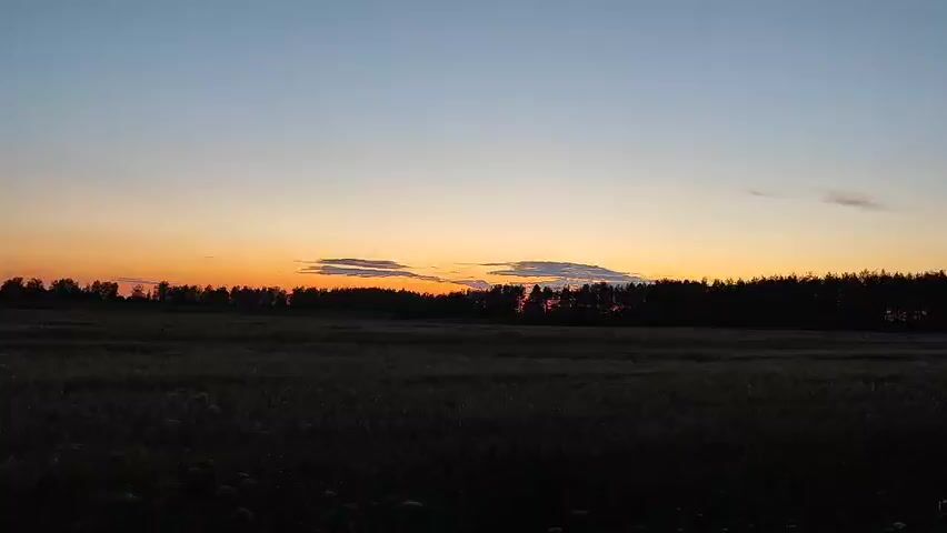 A time-lapse video captures a sunset over a field and a treeline. The sky transitions from a deep blue to a gradient of orange and yellow near the horizon. Silhouetted trees form a dark band across the middle of the frame, with a dark field of vegetation in the foreground. The scene is peaceful and serene.