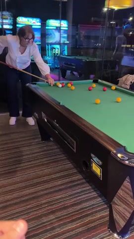 A woman wearing a white blouse, dark pants, and white shoes lines up a shot on a billiard table. She stands to the left of the table, facing it, and holds a pool cue. The billiard table has a green felt surface and multiple yellow and red balls are dispersed across it. The table has a dark brown wooden frame with an "Atlantic" brand label. The floor is covered in a multicolored striped carpet. Behind the table, large glass windows reflect the interior of an arcade with bright neon lights. Another pool table is visible beyond the glass. A man's foot is visible in the lower left corner of the frame. She hits the cue ball, then steps away from the table.