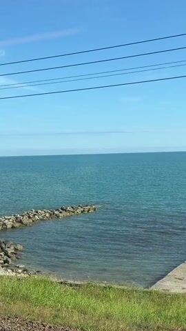 This is a vertical video shot from a moving vehicle. It shows a body of water with a clear blue sky above. In the foreground, there is a strip of green grass. A concrete pier juts out into the water, and on it, four people are seated. To the left, a rocky breakwater extends into the water. In the distance, overhead power lines are visible against the sky. As the video progresses, a utility pole with some equipment attached to it appears on the right side of the frame. The vehicle continues to move, and the pier with the seated people recedes into the background.
