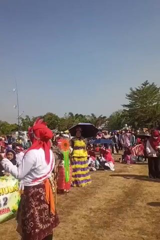 The video shows an outdoor event, likely a parade or festival, under a clear blue sky. In the foreground, a person wearing a red headscarf, white shirt, and a patterned skirt stands facing away from the camera, holding a banner. The banner, visible on the left, has text including "MI NURUL HUDA KELAS 2 CERDAS CERIA". To the right of this person, another individual is dressed in a colorful yellow and purple costume with a tall, elaborate headdress. A black umbrella is held above them. A crowd of people, many wearing headscarves and colorful clothing, are gathered in the background, some sitting on the ground and others standing. In the distance, trees and structures are visible. The ground is covered in dry grass and dirt. There are several kites or similar objects flying in the sky, attached to long strings. The overall atmosphere appears festive and lively.