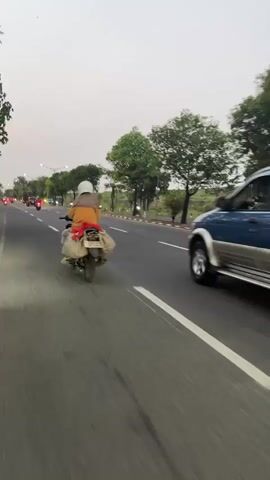 The video shows a side view of a road with multiple vehicles moving in the same direction. In the foreground, a motorcycle is traveling on the left lane, followed by a blue SUV in the right lane. The SUV has a spare tire mounted on the rear. Further ahead, a white car is visible, and then a large tanker truck with colorful decals. Other motorcycles are also seen on the road. The environment appears to be an open road with trees lining the sides and a cloudy sky in the background. The video seems to be shot from another moving vehicle.