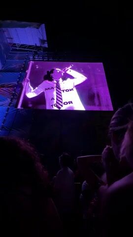The video captures a concert scene from the perspective of an audience member. In the foreground, the silhouettes of several people are visible, their faces and upper bodies obscured by darkness. The focus is on a large screen above the stage, which displays a performance. On the screen, an individual, possibly a performer, is illuminated by purple lighting. They are wearing a white shirt with a tie and have dark hair styled in pigtails. Text is visible on the shirt. The screen is framed by stage lighting and scaffolding on the left side. The overall atmosphere suggests a nighttime event with a significant crowd.