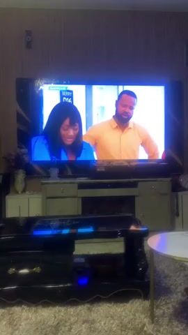 The video captures a panoramic view of a living room. A large flat-screen television is centered, displaying a show with two individuals and English subtitles reading, "You know what, Inyiye, it's okay." Below the TV is a multi-tiered entertainment console. To the left, a large speaker cabinet is visible. The camera pans right, revealing a person's lower legs and feet, clad in dark trousers, resting on a sofa. A small round white side table is beside them, and a laptop is open on the sofa cushion. In the foreground, a dark rectangular coffee table with gold accents sits on a brown shag rug. An ornate gold and white sofa is visible further right. The background features a patterned wall, a white vertical bookshelf with decor, a wall clock, and a white air conditioning unit.