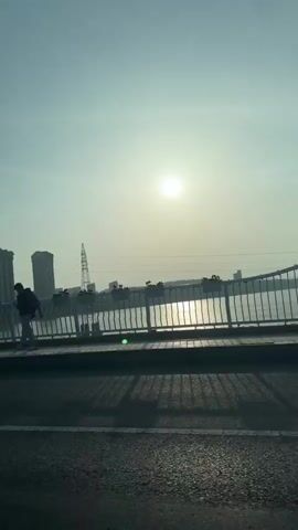 The video shows a view from a vehicle driving across a bridge on a sunny day. The sun is bright and reflects off the water below. On the bridge sidewalk, a person stands looking out at the water. Buildings and a power line are visible in the distance. There are guardrails along the side of the bridge.