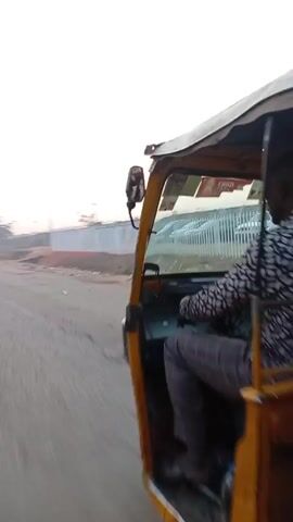 The video shows a view from inside a moving vehicle, likely a tuk-tuk, on an asphalt road. The sun is rising in the background, creating a hazy sky. A man is walking alongside the road. A fence lines the side of the road, and utility poles with overhead wires are visible. The overall environment appears to be a suburban or peri-urban area.