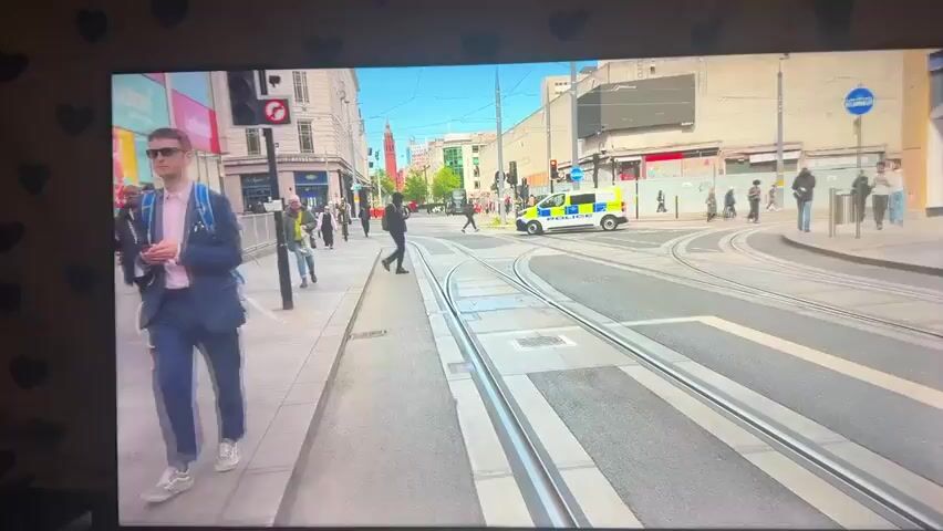 The video opens on a busy city street with trams running through it. People are walking on the sidewalks, and a police car is visible in the distance. The perspective shifts to a street with parked cars and pedestrians on the sidewalk. The camera is positioned low, possibly on a bicycle, as evidenced by the handlebars in the foreground. The view then transitions to a street with two police vehicles parked, one with its rear illuminated. Construction barriers are visible on the right side of the road. The scene depicts an urban environment with everyday activities and a police presence.