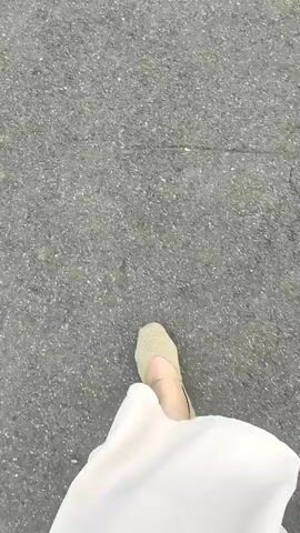 The video shows a person's feet walking on a paved surface. The person is wearing light-colored shoes and a long white garment that obscures their legs. The camera is positioned at a low angle, looking down at the ground as the person walks away from the camera. The paved surface appears to be asphalt. There are no other people or objects visible in the video. The lighting suggests it is daytime. The action is simply walking.