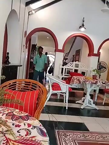 The video shows the interior of a home, probably a set for filming. The room has three arched doorways leading to other rooms. The walls are white with red trim around the arches. There is black and white checkered flooring with decorative rugs. Furniture includes white wicker chairs with red and white striped cushions, a white round table, and a rattan chair with a colorful cushion. A man in a light blue shirt and green pants walks across the room. Other people can be seen in the background, through the archways. A ceiling fan and a standing fan are also visible.