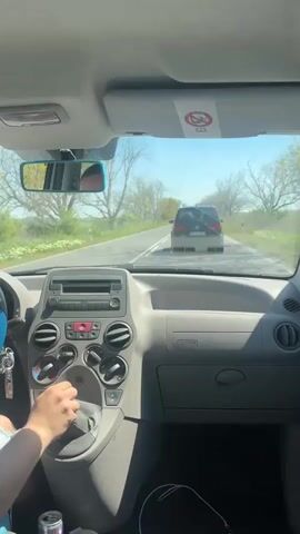 The video shows a car's interior from the driver's perspective, focusing on the dashboard and the road ahead. A person's hand is visible operating the controls of the car's ventilation or climate system. The car is moving on a two-lane road, following another vehicle. The sky is clear, and trees line the sides of the road. Another car passes in the opposite direction.