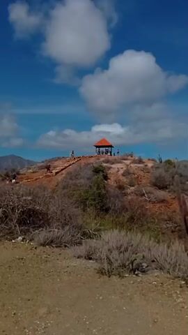 A wide shot captures a arid, rocky hilltop under a bright blue sky with scattered white clouds. A small, open-air pavilion with an orange roof stands at the summit, surrounded by a few people. A dirt path with stone steps winds its way up the hill, and several individuals are visible walking on it and around the pavilion. The landscape is characterized by sparse, dry vegetation and various types of cacti. The camera pans across the scene, revealing the ocean in the distance and more cacti and dry brush in the foreground. The video shows a man in a blue shirt and shorts walking, followed by two women walking on a dirt path. The woman in front is wearing a short, light-colored dress, and the woman behind her is wearing a red jumpsuit. They are walking towards the camera. The scene is set in a sunny, outdoor environment.