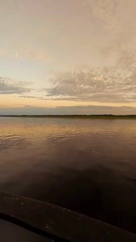 The video shows a wide shot of a calm river at sunset. The sky is filled with scattered clouds tinged with hues of orange and yellow from the setting sun. The horizon line is marked by a strip of land covered in green vegetation. The water reflects the colors of the sky, creating a serene and peaceful atmosphere. The lower portion of the video shows a dark, blurry foreground, possibly the edge of a boat or dock. There are no people or animals visible in the video. The video appears to be filmed from a stationary position.