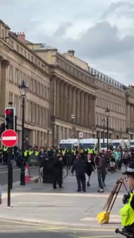 The video shows a street scene with a large group of police officers in yellow vests forming a line in front of multiple white police vans. Behind the police line, there are more police officers. Several people are walking on the sidewalk and crossing the street. A traffic light shows a red light and a no entry sign. There are also street lamps and a ladder in the foreground. The background features tall buildings with a classical architectural style. The sky is overcast.