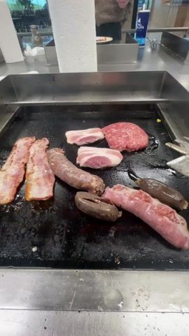 The camera shows a cooktop with various meats being cooked. The cooktop is black and greasy with several slices of bacon on the left, two pieces of pork belly and a raw hamburger patty in the upper center, and several sausages of different colors in the lower center. In the background, a person in a brown sweater is visible, using tongs to manipulate the meats. There is a roll of paper towels and a soda cup on the counter behind the cooktop. The environment appears to be a commercial kitchen or food service area.