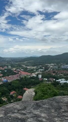 This is a vertical video shot from a high vantage point, looking out over a sprawling city nestled among rolling green hills. The foreground is dominated by a rough, grey rock formation. Beyond the rock, the city unfolds with a patchwork of red-tiled roofs, modern buildings with white and colored facades, and numerous trees. In the distance, more hills and mountains rise, forming a natural backdrop to the urban landscape. The sky is a vibrant blue, dotted with fluffy white clouds, suggesting a pleasant day. The video pans slowly to the right, offering a panoramic view of the city and its surroundings.