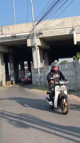 The video shows a person wearing a black hoodie and a helmet riding a white scooter on a paved road. The scooter is moving towards the viewer. In the background, another person is riding a red cart. The scene is outdoors with a concrete overpass above the road. Overhead power lines are visible. The environment appears to be a sunny day.