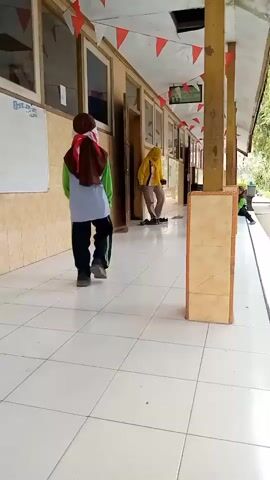 Two individuals, likely students based on their attire and the environment, are observed in a school hallway. The hallway features tiled flooring, beige walls with windows, and a covered walkway supported by wooden pillars. Red and white triangular flags are strung along the ceiling. One individual, wearing a red hijab and a green and grey outfit, walks away from the camera. Another individual, wearing a yellow jacket and dark pants, is further down the hallway and appears to be sweeping the floor. The camera perspective is low and positioned on the tiled floor, looking down the hallway. The lighting suggests it is daytime, with natural light entering from the right side of the frame.