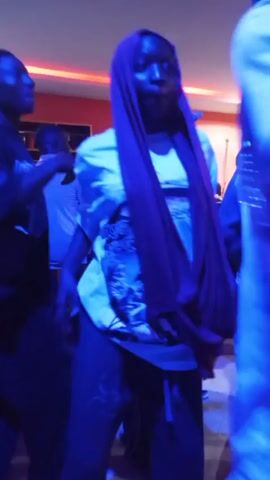 The video captures a group of people in what appears to be a dimly lit indoor space, possibly a bar or club, with neon-style lighting casting a strong blue and pink hue. The central figure is a person wearing a white t-shirt and a vibrant purple headscarf, with their hair also appearing to be purple. They are looking down and seem to be singing or speaking, with their hands in motion. Other individuals are visible in the background, some partially obscured by the lighting and the central figure. One person to the left wears a dark shirt and looks towards the center. Another person in the background wears a baseball cap. The overall atmosphere suggests a social gathering or performance.