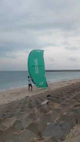 The video captures a serene coastal scene in Phú Quý Special Administrative Region, Vietnam. The main subject is a man in a white t-shirt and dark shorts, standing on a sandy beach. He is holding and controlling a large, vibrant green kite, which bears the word "GONG" prominently on its surface. The kite is inflated and airborne, suggesting windy conditions, possibly indicating preparation for kitesurfing or kiteboarding.

In the background, a second person is visible walking towards the ocean, further into the water. The beach itself features soft sand meeting the gentle waves of the sea. In the immediate foreground, large, grey, concrete interlocking blocks, possibly forming a sea wall or breakwater, line the edge of the beach.

The sky is overcast with a mix of light and dark clouds, suggesting a cloudy day, possibly late afternoon or early morning. The ocean appears calm with subtle waves breaking on the shore. The overall atmosphere is tranquil, focusing on the individual engaging with the kite against the backdrop of the sea and sky.