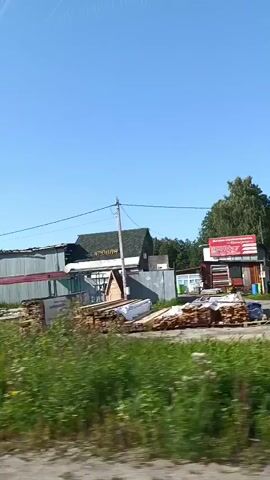 The video shows a view from a moving vehicle on a sunny day. The scene is rural or semi-rural, with a clear blue sky. The foreground consists of tall green grass and weeds. Behind the vegetation, there are various structures. One structure is a long, grey, corrugated metal building with red trim. Further to the right, there is a smaller building with a dark green roof and white walls, and what appears to be a construction or agricultural area with wooden elements. A pedestrian crossing sign is visible on a pole. There is also a blue bus stop shelter with a pink and blue roof. The camera is moving horizontally, capturing the passing scenery.