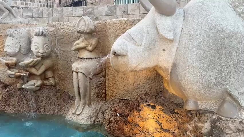 The video shows a fountain with various stone sculptures. In the foreground, a large sculpture of a bull's head is visible on the right, with water spraying from its nostril. To the left, there are sculptures of two child-like figures holding bowls, with water flowing from them. Between these and the bull, a sculpture of a seated child with arms crossed is positioned, also with water spraying from its mouth. Further left, two dolphin-like sculptures are submerged in the blue water of the fountain basin. In the background, more stone sculptures are arranged in tiers, including figures of people and animals. Some of the sculptures are lit, casting warm light on the scene. The environment appears to be an outdoor public space with stone walls and greenery in the background. The lighting suggests it might be dusk or evening.