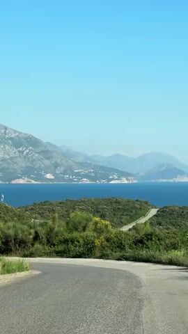 The video shows a scenic view of a mountainous landscape with a blue sea and sky. The camera is mounted on a vehicle moving along an asphalt road, lined with green bushes and some yellow flowers. The mountains in the background appear hazy and distant. There are some small settlements visible along the coastline. The sky is clear blue. The scene is captured in daylight.