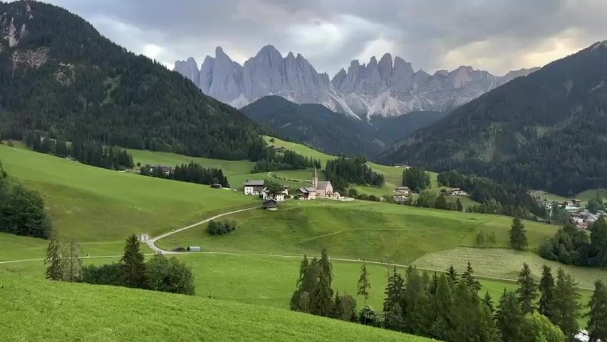 This media file showcases a picturesque landscape in St. Magdalena, Santa Maddalena, Italy. The scene is a rolling green valley nestled between imposing, jagged mountains in the background. The mountains, with their sharp, rocky peaks, are a prominent feature, some with patches of snow or bare rock.

In the mid-ground, a charming village is visible. The focal point of the village is a church with a tall, slender steeple. Several other buildings, likely houses and farm structures, are scattered around the church and along a winding road that traverses the valley. The green hillsides are a mix of manicured pastures and denser evergreen forests.

The time of day appears to be late afternoon or early evening, given the soft, diffused light and the muted colors. The sky is overcast with dramatic, textured clouds, suggesting the possibility of impending rain or a recent shower, contributing to a sense of serene, almost moody, beauty. The overall atmosphere is tranquil and idyllic, evoking a feeling of peace and connection with nature. No people are visible in the scene, emphasizing the solitude and grandeur of the natural environment. The focus is entirely on the harmonious interplay of the built environment and the majestic alpine landscape. There is no visible text in the media file.