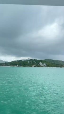 The video shows a boat moving away from an island under a cloudy sky. The island is covered in green vegetation and has some buildings near the beach. The water is turquoise in color, and the boat leaves a white wake behind it. The camera appears to be mounted on the boat.