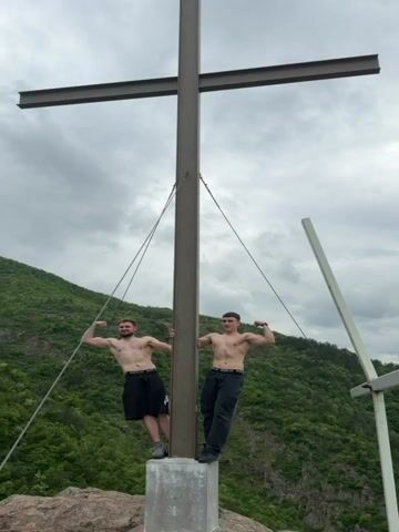 Two shirtless men stand atop a rock formation beside a tall metal cross. They flex their arm muscles. The scenery behind them includes a lush green mountain range, a wide flat valley with a small town, and a cloudy sky. The camera zooms out to show more of the valley view.
