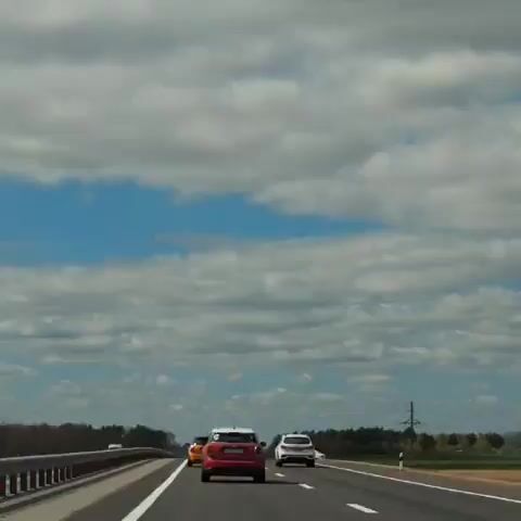 The video captures a daytime view from a vehicle driving on a multi-lane highway in what appears to be Belarus. The sky is bright blue with numerous fluffy white clouds. Lush green fields and a treeline stretch along both sides of the road, with a power line tower visible on the right in the distance.

Several vehicles are present on the highway. Directly ahead of the camera car is a red SUV. To its left, a yellow car is partially visible, while a white SUV drives to the right of the red SUV. Further down the right shoulder, another white car is parked. A white van can also be seen partially on the far left, behind the guardrail. The cars are in motion, driving along the highway. No discernible text is visible on any of the vehicles or road signs.