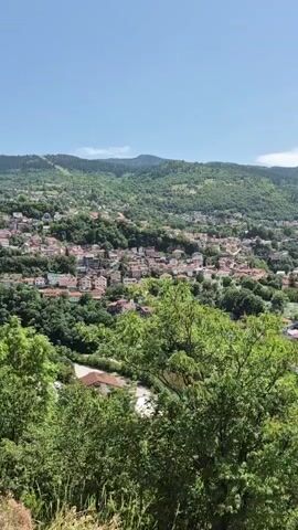 A drone shot pans over a city nestled in a valley surrounded by lush green hills. The city features a dense collection of buildings with red tile roofs, interspersed with trees. A winding road is visible, with a black car driving on it. In the foreground, the branches of green trees obscure parts of the view. The sky is blue with some scattered clouds. A body of water, possibly a lake or river, is visible on the left side of the frame. The overall impression is of a scenic, urban landscape in a hilly, vegetated environment.