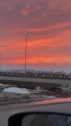 The video shows a roadside view of a highway during a vivid sunrise or sunset. The sky is a blend of red, orange, and pink hues, with some wispy clouds scattered across the expanse. Along the highway, cars and a semi-truck are seen driving past, and a long train with various tanker cars sits on tracks in the distance. There are streetlights lining the road, and patches of melting snow are visible on the ground beside the highway. The video seems to be taken from inside a car, with the dashboard visible in the foreground.