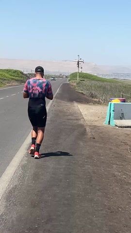 A man is walking along a paved path next to a two-lane road on a clear day. He is wearing a colorful top, black shorts, red running shoes, black socks, and a black baseball cap. The road runs parallel to the path. Beyond the road is vegetation. Across the road, there is a painted concrete barrier with a yellow, blue, and white design. In the distance, there are hills. A car passes on the road.