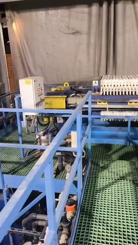 The video shows an industrial setting with large machinery and a textured green floor. In the foreground, a blue metal railing and walkway overlook the machinery. The machinery itself is blue and white with various pipes, wires, and control panels visible. A large white tank with a logo is positioned to the right of the machinery. The background features a gray curtain and some overhead lighting. The overall impression is of an active industrial processing area.