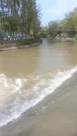 The video shows a wide, muddy river with a small man-made waterfall or dam creating whitewater. On the left bank of the river, there is a paved area with parked cars and trees. Several people are visible walking along the bank. A metal bridge crosses the river in the background, with a few people also visible on the bridge. The right bank of the river is lined with trees. The sky is partly cloudy. The overall impression is of a public park or recreational area along a river.