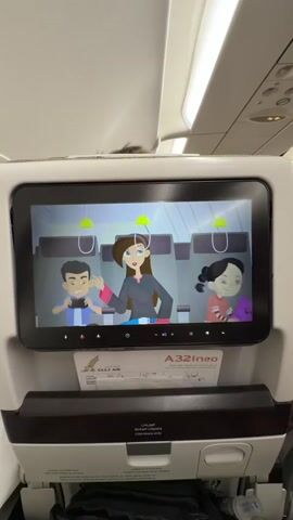 Here's a description of the video:

The video shows an airplane seatback entertainment screen. The screen is displaying a cartoon safety video. The cartoon features a family of three: a boy, a woman, and a girl. They are demonstrating how to use the oxygen masks that drop from the ceiling in the event of an emergency. The cartoon is colorful and clear. The screen is mounted in the seatback of a Gulf Air A321neo aircraft. Below the screen, there is a literature pocket and other seat components.