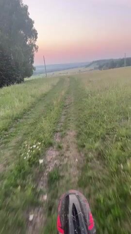 The video shows a first-person perspective of someone riding a dirt bike down a narrow dirt path through a grassy field at sunset. The sky is a soft gradient of pink, orange, and pale blue. Trees are visible on the left side of the frame, and utility poles are scattered across the landscape. The handlebars of the dirt bike, with the "DAREX RACING TEAM" logo, and a digital display are visible in the foreground. The camera is slightly shaky, indicating movement.