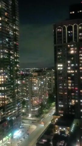 The video captures a nighttime view of a city skyline, dominated by tall buildings illuminated by numerous windows. The camera pans slightly, revealing a street with a few cars driving by. The sky is dark with some clouds, and the distant city lights create a hazy glow. The overall atmosphere is one of urban nighttime activity.