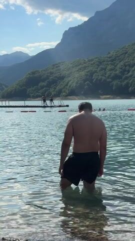 A person stands waist-deep in clear, turquoise water at the edge of a lake. They are facing away from the camera, looking towards the open water. In the distance, several people are visible swimming and playing in the lake. A floating platform with red buoys is positioned in the water. Lush green mountains covered in trees form the backdrop, with a clear blue sky overhead and sunlight illuminating the scene. The water is calm, with ripples reflecting the light.