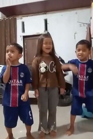 Three young children, two boys and one girl, are standing and singing in a room. The two boys are wearing identical blue and red soccer jerseys and shorts. The girl is wearing a brown long-sleeved shirt and grey pants. The background appears to be a living room with a motorcycle, curtains, and a wall with a window. The children are looking at the camera and appear to be performing a song or dance. The video is in portrait mode.