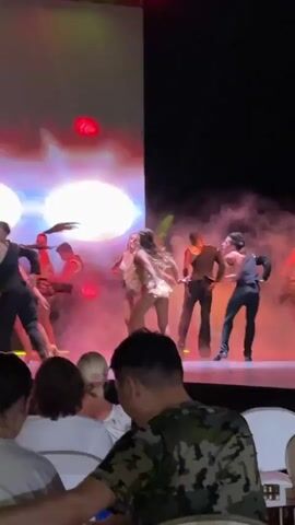 A group of performers are on a stage.  In the foreground, a woman in a sparkling gold costume is center stage, dancing.  Surrounding her are other performers in various costumes, some in black and gold outfits, others in shimmering or leotard-style costumes.  A large screen behind the performers displays abstract, glowing imagery in shades of pink, white, and red.  Smoke or fog machines are in use, creating a hazy atmosphere.  The stage is lit with colorful stage lights, predominantly red and yellow.  In the lower foreground, out of focus, are members of an audience seated in chairs.