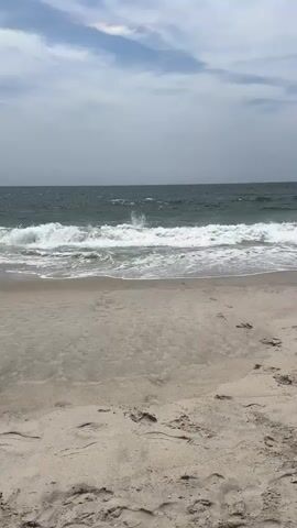 The video shows a view of the ocean and a sandy beach. The waves are crashing on the shore, and there are some clouds in the sky. The sand has footprints on it, indicating that people have been there. The overall scene is calm and peaceful, suggesting a day at the beach.