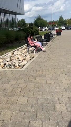 Two young girls are sitting on a park bench outside. The girl on the left is wearing a pink tracksuit and the girl on the right is wearing a white t-shirt and light purple pants. They are both looking away from the camera. The bench is black and made of metal. The area around the bench is paved with grey brick. To the left of the bench is a rock garden with a variety of stones and some green plants. Behind the rock garden is a modern building with large windows. In the background, there is a parking lot with several cars and some trees. The sky is cloudy.