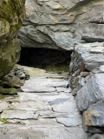 The video shows a low, dark cave entrance at the top of a set of stone steps. The walls around the steps and cave entrance are made of large, gray stones. Some moss grows on the stones. The cave's interior is dark and filled with smaller stones.